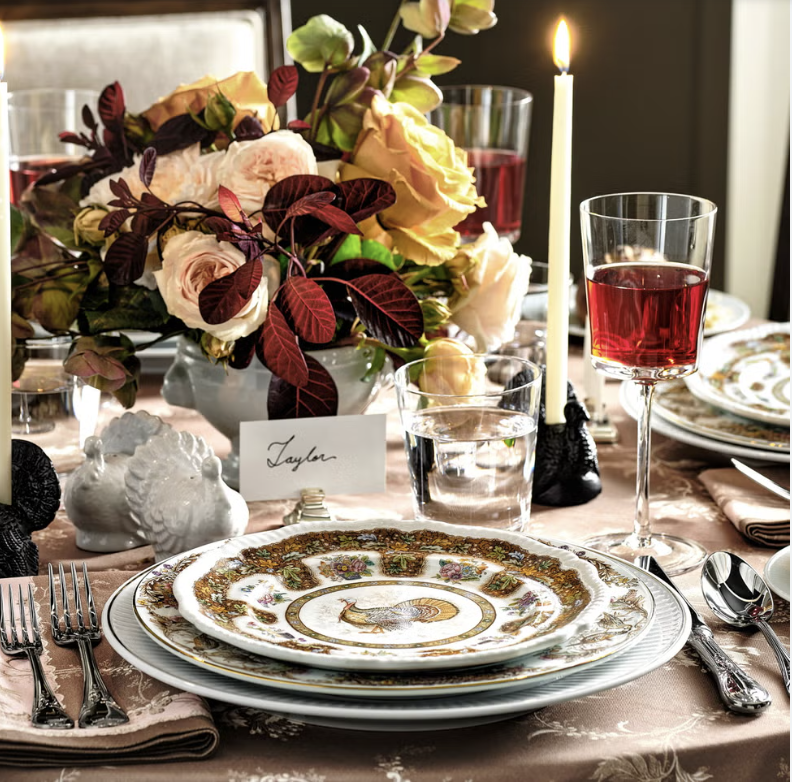 Beautifully intricate thanksgiving table scape with layered plates, flowers, red wine, and tapered candle sticks